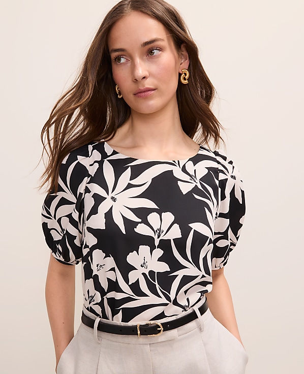 A woman in a black and white floral blouse and light-colored pants stands against a plain, light-colored background.