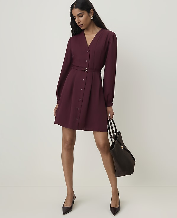A woman in a burgundy dress and heels stands with a tote bag against a plain, light background.