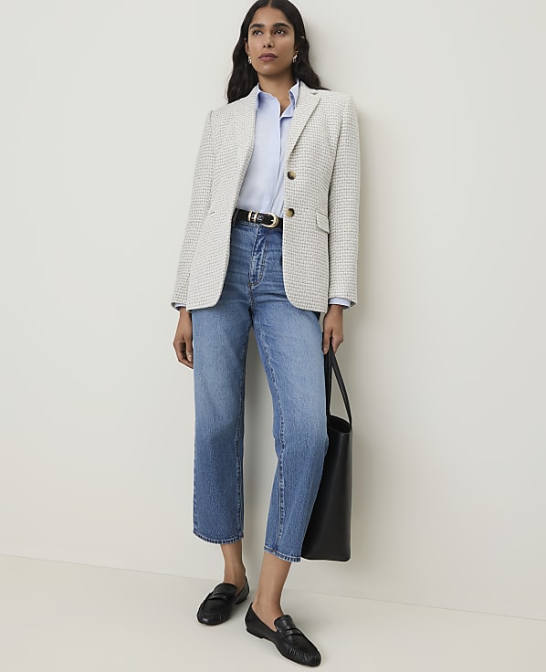 A woman in a light-colored tweed blazer, blue shirt, and jeans stands against a plain white wall.