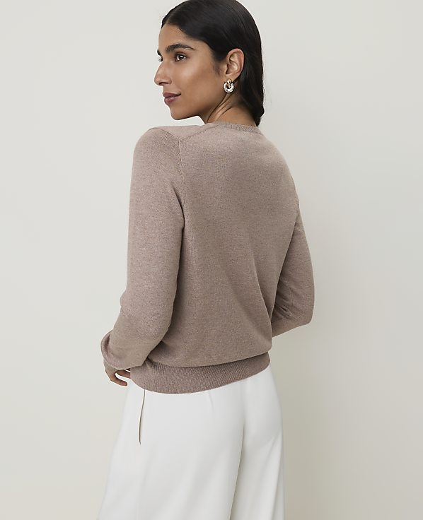 A woman in a taupe sweater and white pants stands against a plain, light background.