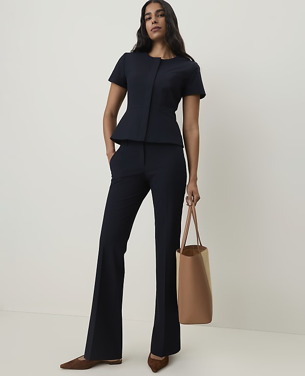A woman in a navy peplum jacket and flared trousers stands with a tan tote bag against a plain, light background.