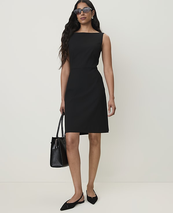 A woman in a black dress and flats stands holding a black handbag against a plain white background.