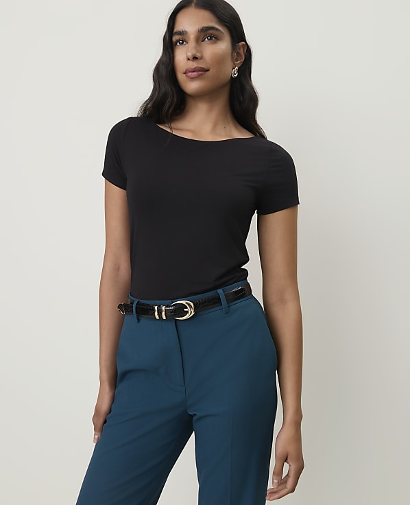 A woman in a black boatneck shirt and teal pants stands against a plain white background.