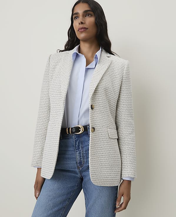 A woman in a light blue shirt, blue jeans, and a textured white blazer stands against a plain white background.