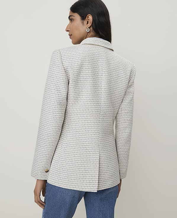 A woman in a tweed blazer and jeans stands against a plain, light-colored background.