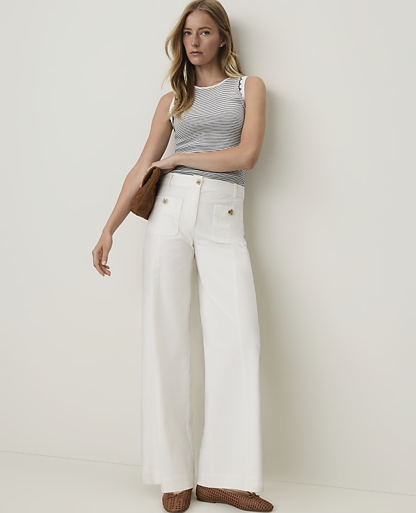 A woman in a striped top and white wide-leg pants stands against a plain white background, holding a brown clutch.