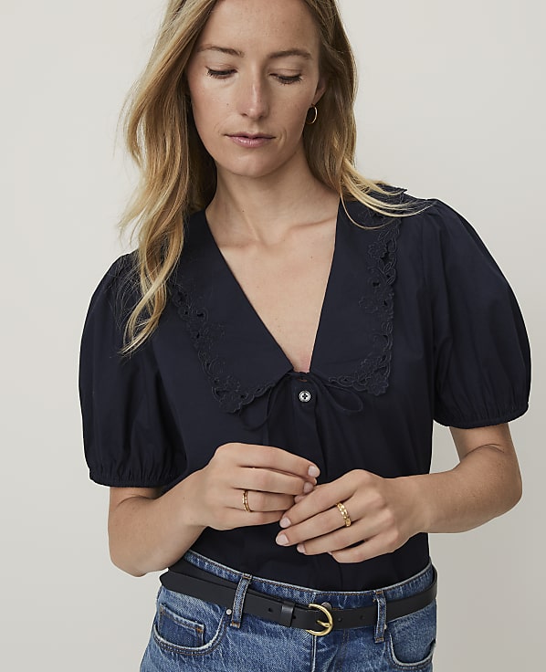 A woman in a navy blouse with a large collar and jeans stands against a plain white background.