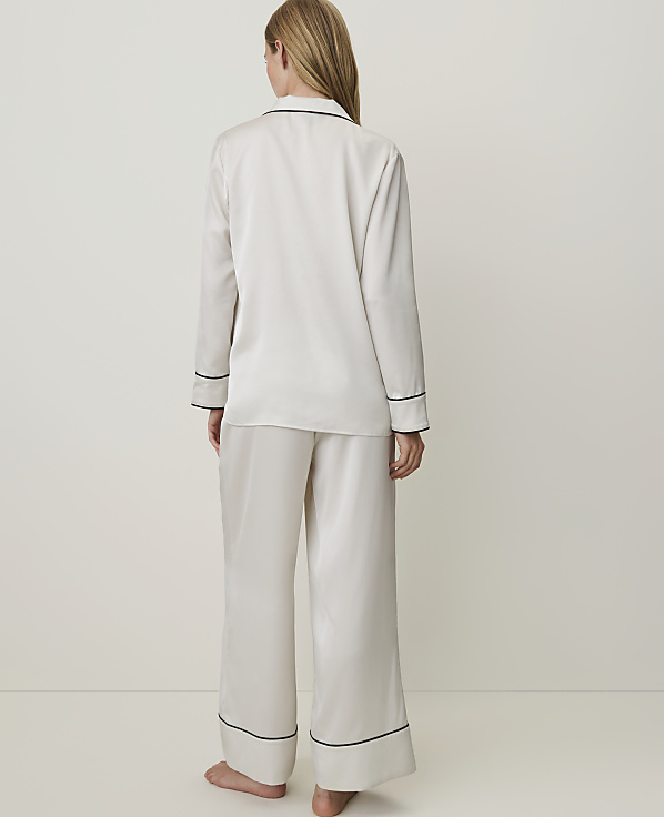 A woman in cream silk pajamas with black trim stands against a plain white background.
