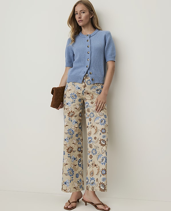 A woman in a blue cardigan and floral pants stands against a plain background, holding a brown clutch.