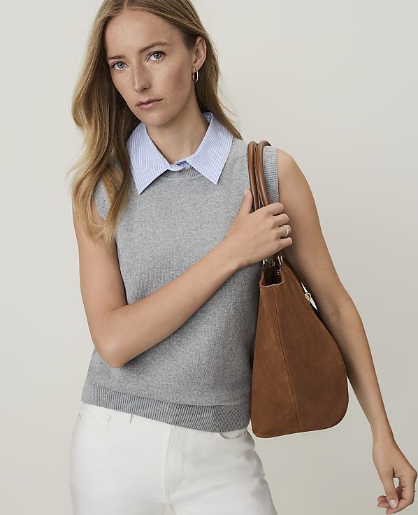 A woman in a gray sweater vest and blue collared shirt holds a brown suede purse against a plain, light background.