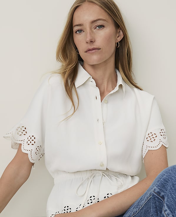 A woman in a white eyelet shirt and blue jeans sits against a plain, light-colored background.
