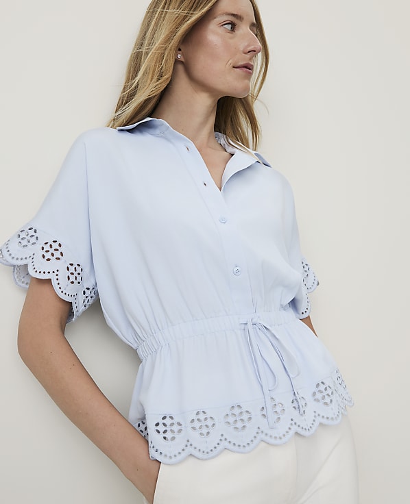 A woman in a light blue eyelet-trimmed blouse and white pants stands against a plain, light background.