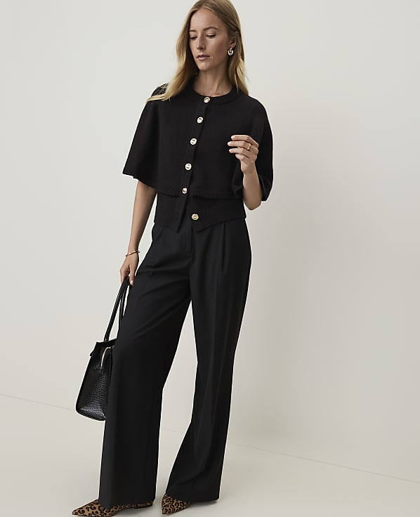 A woman in a black cardigan and wide-leg pants stands against a plain white background, holding a black handbag.