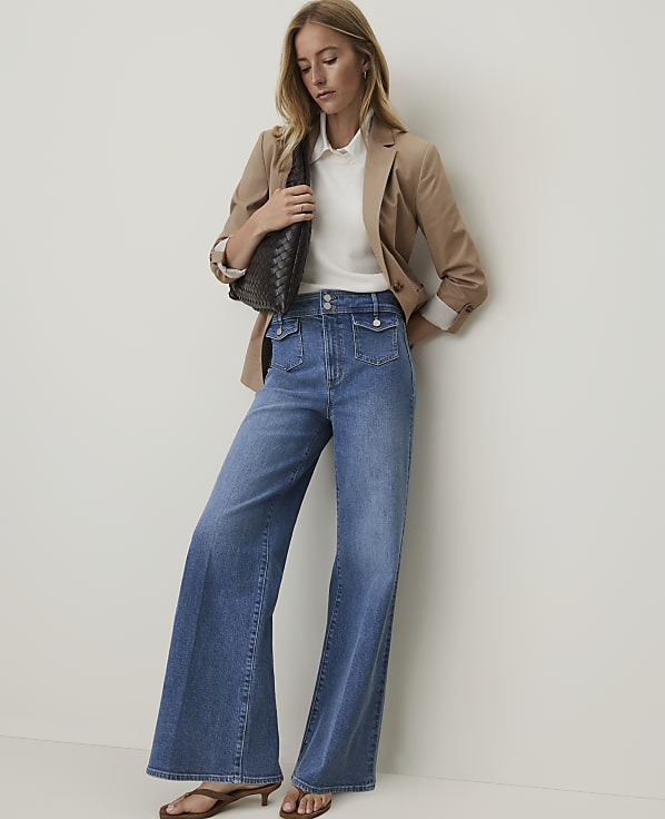 A woman in wide-leg jeans and a blazer stands against a plain white wall.