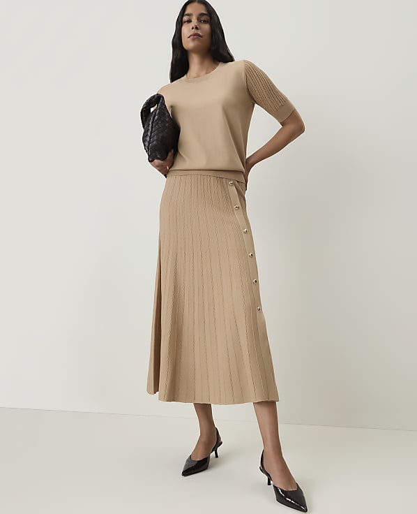 A woman in a beige knit top and pleated skirt stands against a plain, light background, holding a black clutch.