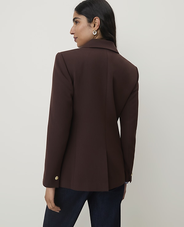 A woman in a brown blazer and dark jeans stands against a plain, light-colored background.