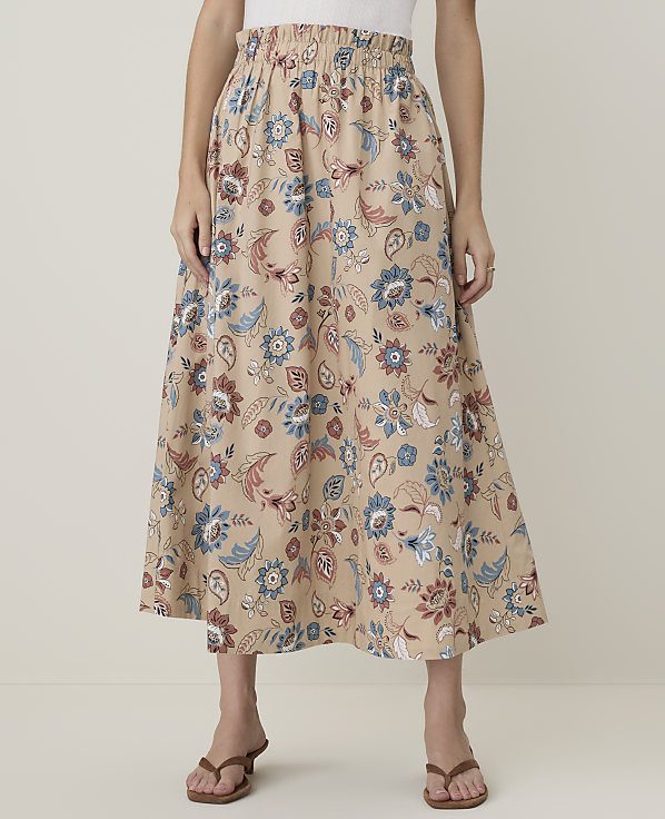 A person stands wearing a beige floral skirt with blue and rust-colored patterns, against a plain, light background.