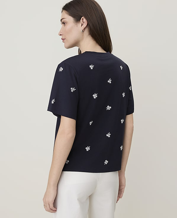 A woman in a dark blue t-shirt with white floral embroidery stands against a plain, light-colored background.
