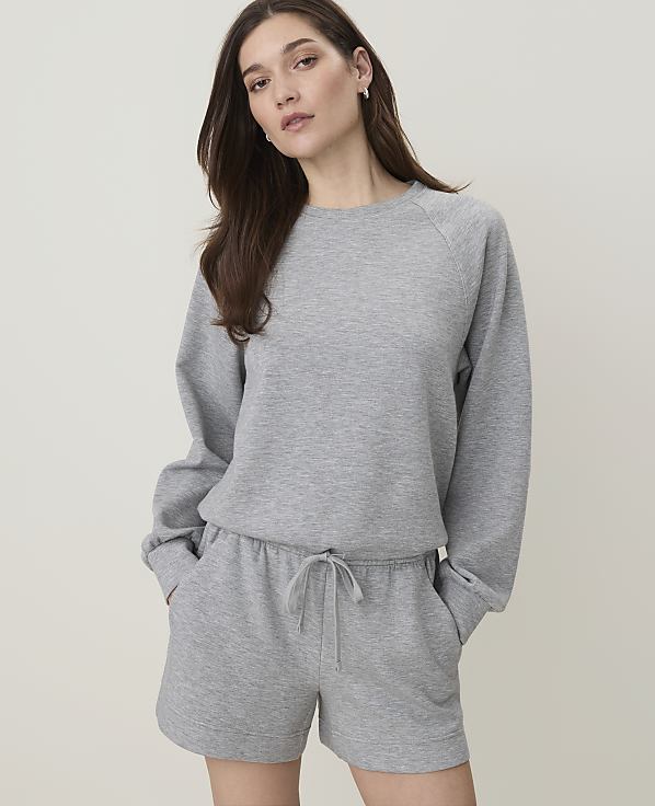 A woman in a grey sweatshirt and shorts stands against a plain, light grey background.