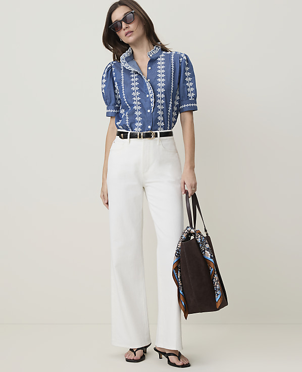 A woman in a blue embroidered shirt and white pants stands against a plain, light-colored background, holding a brown tote bag.