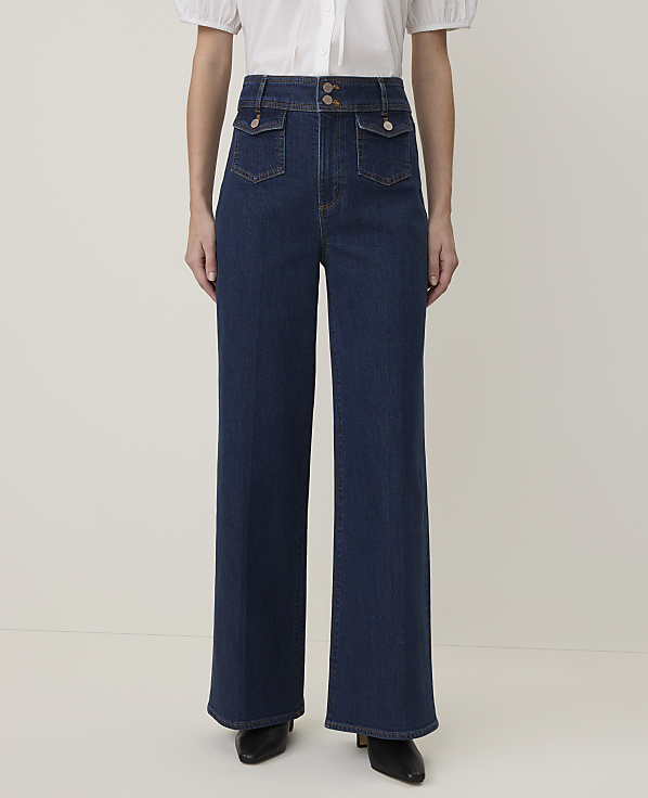A person stands in dark wash wide-leg jeans with a white blouse and black heels against a plain, light background.