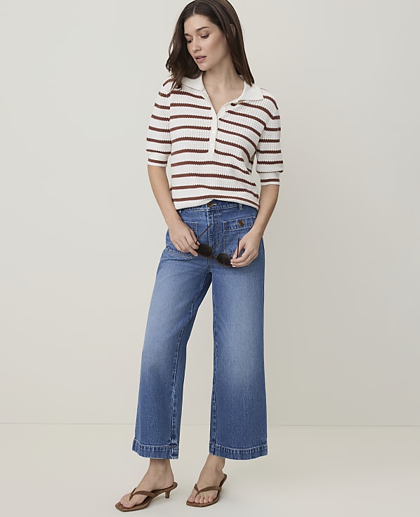A woman in a striped polo shirt and wide-leg jeans stands against a plain, light-colored background.