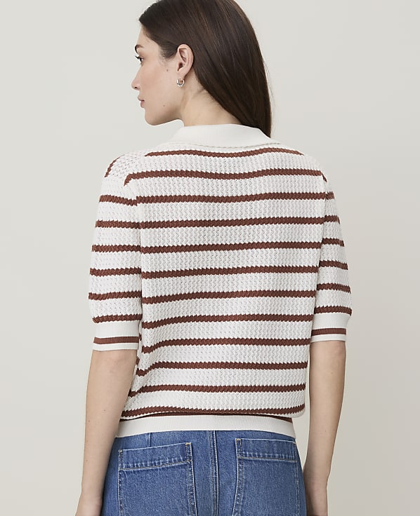 A woman in a striped polo shirt and jeans stands against a plain background.
