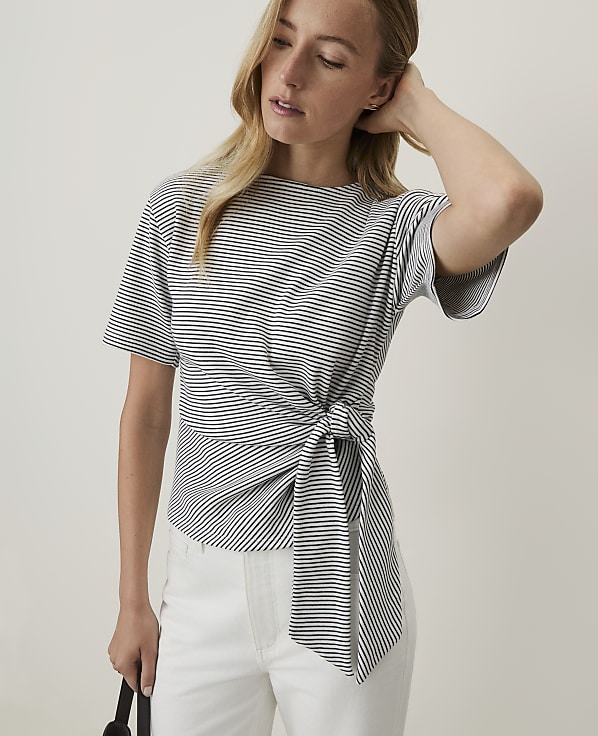 A woman in a striped tie-waist top and white pants stands against a plain, light background.