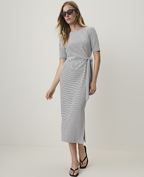 A woman in a grey and white striped dress and black sandals stands against a plain, light-colored wall.