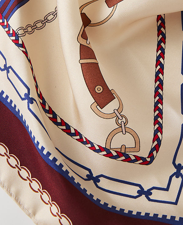 A cream-colored silk scarf with a equestrian-themed print of chains and leather straps is folded in the foreground, with a dark red border visible at the bottom left.