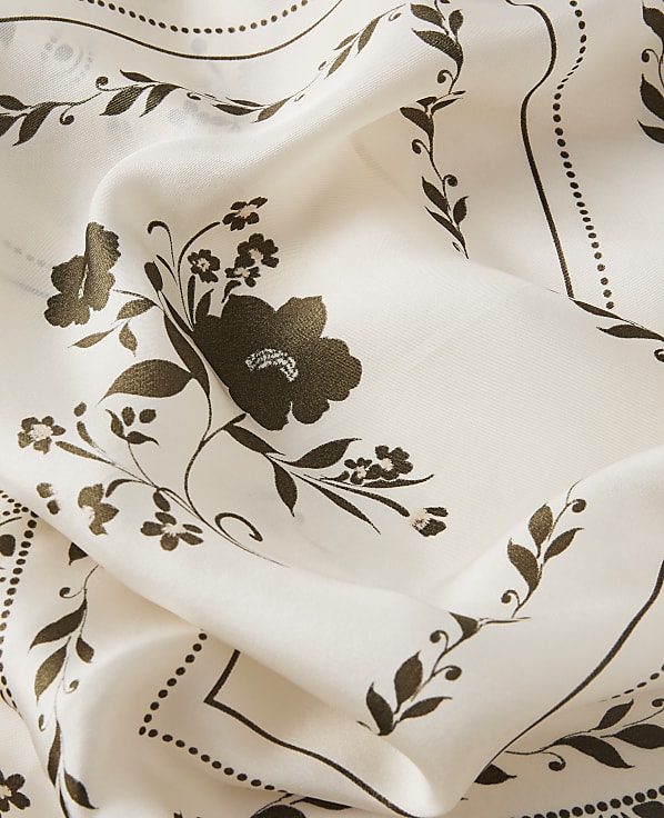 A cream-colored fabric with a delicate floral pattern in dark brown is folded, revealing intricate swirling vines and small blossoms in the foreground and a repeating border design in the background.