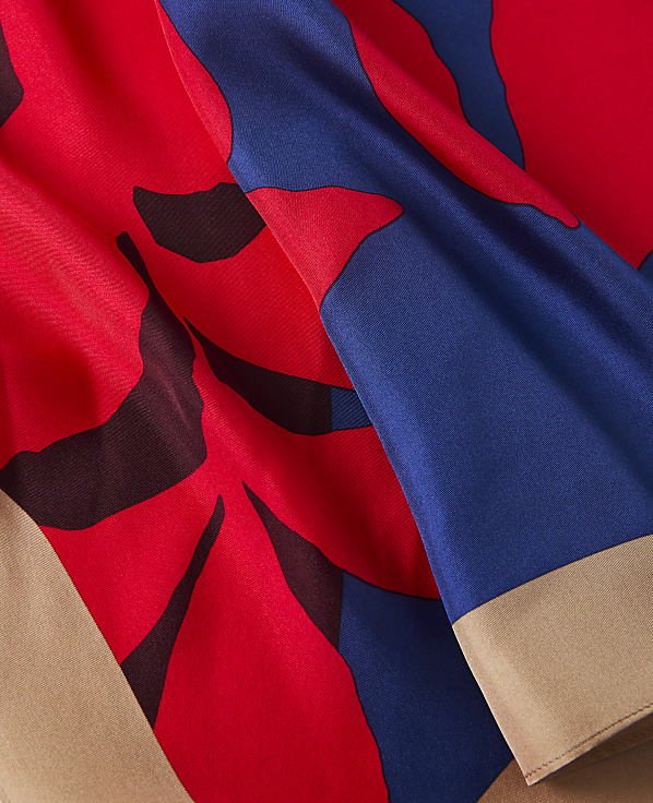 A close-up of a flowing fabric with a bold red and blue floral pattern in the foreground, transitioning to a plain tan border in the background.