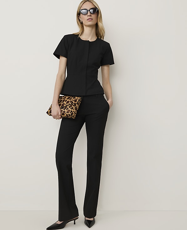 A woman in a black outfit and sunglasses holds a leopard print clutch in front of a plain white background.