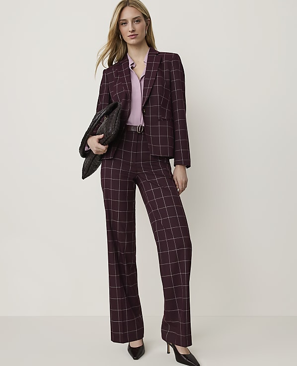 A woman in a burgundy plaid suit and pink blouse stands against a plain, light-colored background, holding a black clutch.