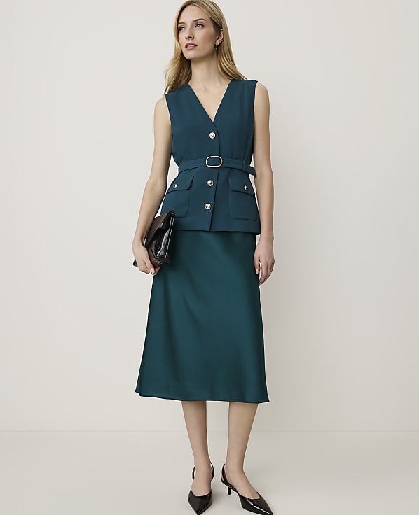 A woman in a teal vest and skirt stands against a plain white background, holding a dark clutch.