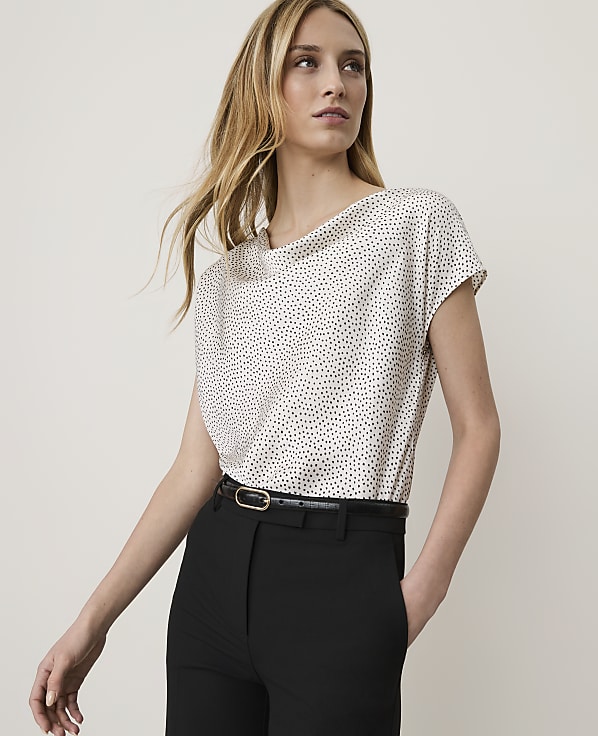 A woman in a polka dot blouse and black pants stands against a plain, light background.