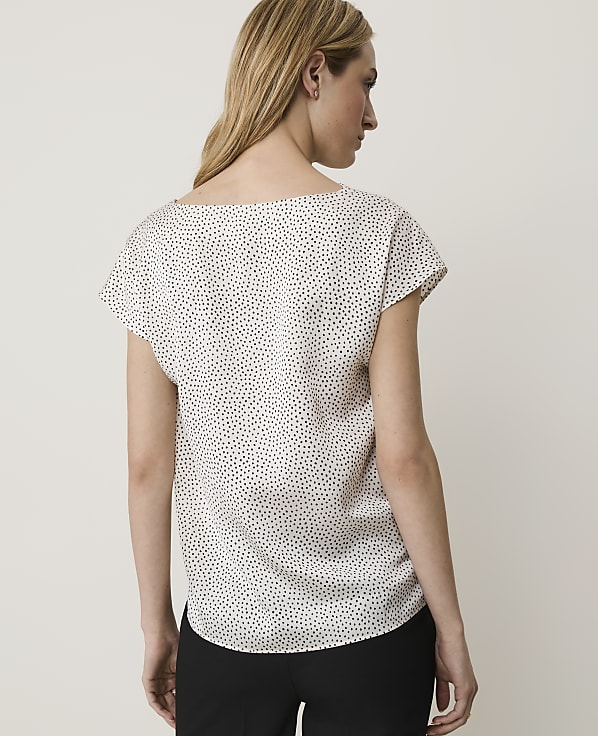 A woman in a white polka-dot blouse stands against a plain, light-colored background.