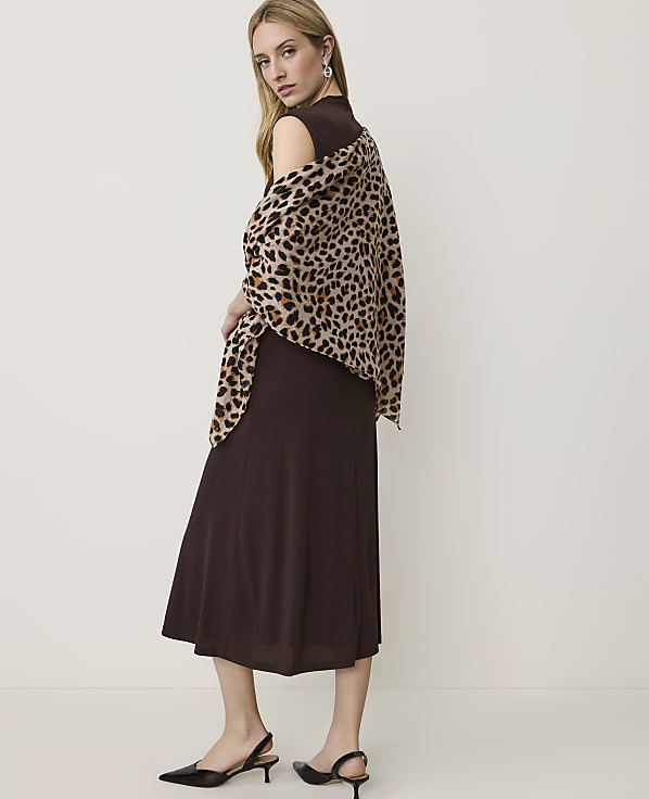 A woman in a brown dress and leopard print shawl stands against a plain white background.