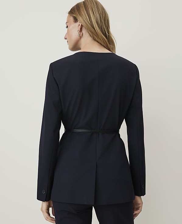A woman in a navy blazer cinched with a black belt stands against a plain, light gray background.