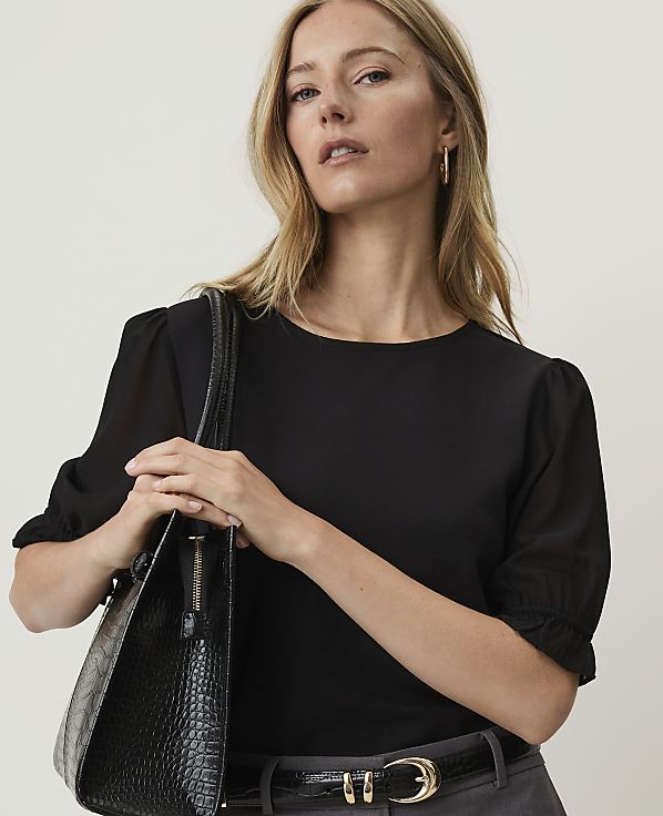A woman in a black top and grey pants holds a black textured handbag against a plain background.