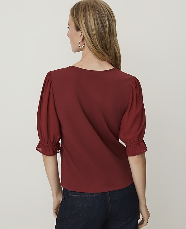 A woman in a maroon top with puffed sleeves and dark jeans stands against a plain white background.
