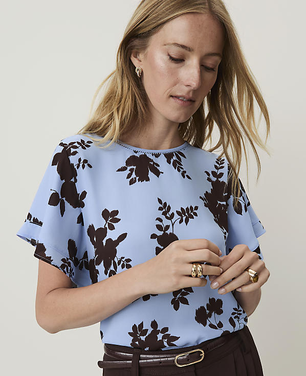 A woman with blonde hair wears a light blue blouse with a dark floral print, while a plain, light-colored background is visible.