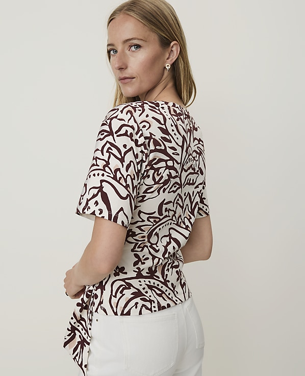 A woman in a patterned top and white pants stands against a plain white background.