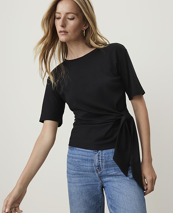 A blonde woman in a black tie-waist top and blue jeans poses against a plain gray background.