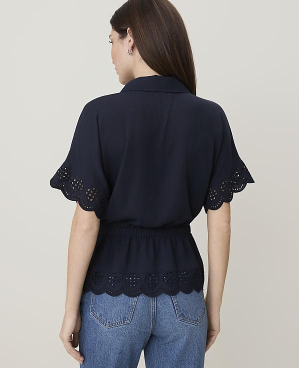 A woman in a navy blue blouse with eyelet trim and blue jeans stands against a plain white background.