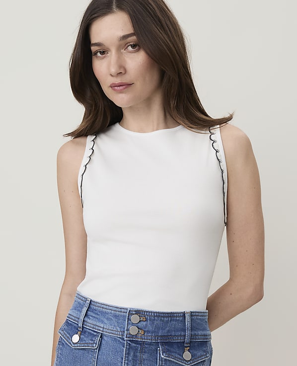 A woman in a white scalloped-edge tank top and blue denim jeans is shown against a plain white background.