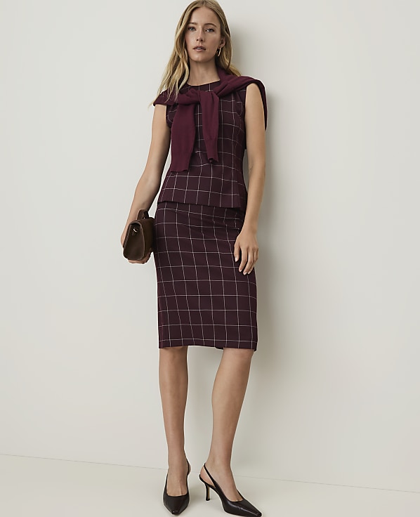 A woman in a burgundy plaid dress and sweater draped over her shoulders stands against a plain white background, holding a brown clutch.