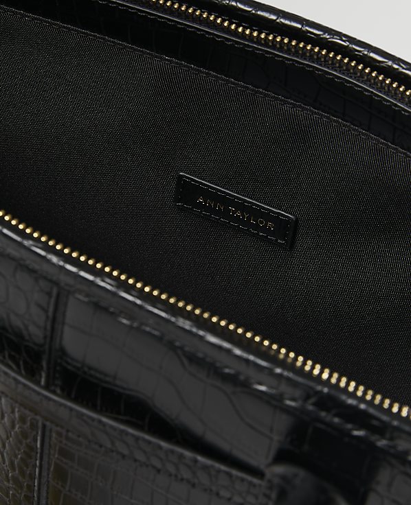 A black crocodile-embossed leather bag with a gold zipper is shown against a dark, textured background with the "Ann Taylor" logo visible inside.