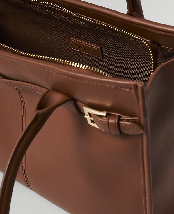 A close-up of a brown leather handbag with a gold buckle in the foreground is set against a blurred, neutral background.