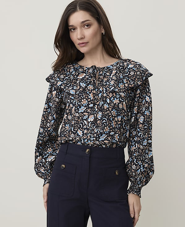 A woman in a floral blouse and dark pants stands against a plain white background.
